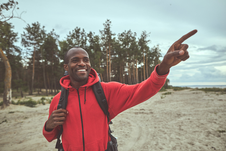 Hiking with pleasure. Waist up portrait of tourist standing near the pine forest showing track directionの写真素材