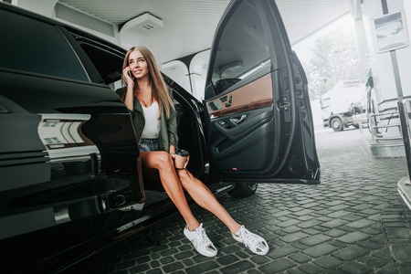 Full length portrait of happy girl taking on mobile while relaxing on seat of modern car. Glad...