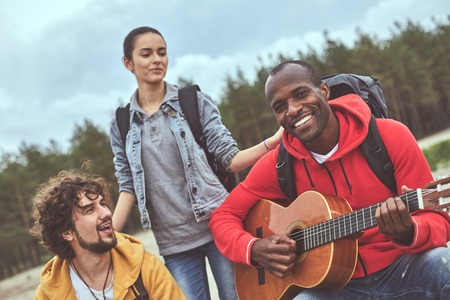 Waist up portrait of two happy man sitting and jamming on guitar while their female friend standing and listeningの写真素材
