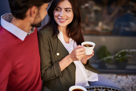 You are the best. Charming young lady with cup of tea sitting at the table and looking at bearded man. Gentleman hugging his smiling lovely girlfriendの写真素材