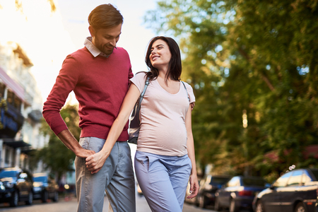 Perfect day with my love. Portrait of smiling young gentleman hugging his beautiful pregnant wife and looking at her tummy. Lady touching husband arm and staring at him with smileの写真素材