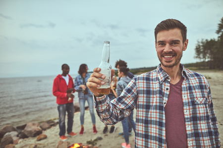 Portrait of smile man holding bottle of beer in hand. His friends standing on background near bonfireの写真素材