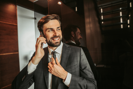Portrait of cheerful unshaven businessman telling on mobile while straightening tie indoorの写真素材