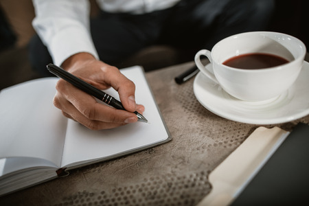Top view close up of male hand holding pen and making notes in his business diary. He is drinking hot beverage in large mugの写真素材