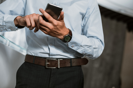 Low angle close-up of male hands using smartphone. Man in office clothes is standing and holding gadget. He is typing on touch screenの写真素材