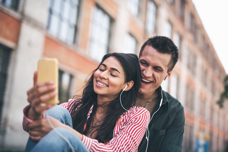 Wonderful song. Expressive young man laughing and looking at the smartphone while his relaxed girlfriend enjoying the music with her eyes closedの写真素材