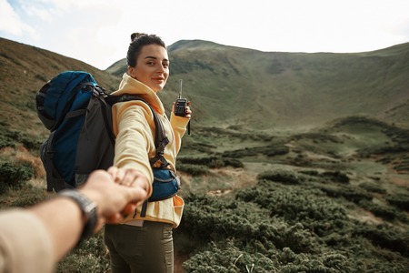 Looking curious. Confident calm young woman looking at you and smiling while being in the mountains with convenient radio set in her handの写真素材