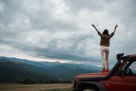 Rear view of a young slim female tourist standing in the car hood while enjoying view over the mountainsの写真素材
