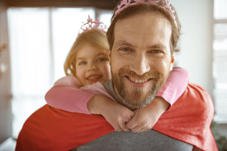 Wonderful moments together. Portrait of happy father carrying girl on shoulders. He is looking at camera and smiling. Man is wearing crown and mantleの写真素材