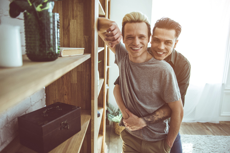 Waist up portrait of happy gay couple cuddling while standing in apartment near wooden wall unit with shelvesの写真素材
