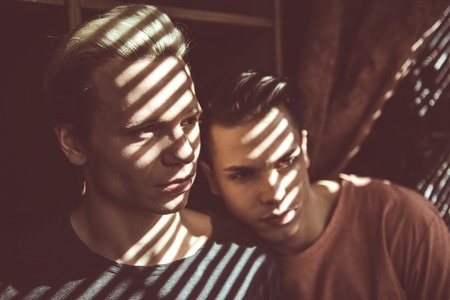 Close up toned portrait of attractive guy with shadow from jalousie on his face looking away with serious expression, while boyfriend laying head on his shoulderの写真素材