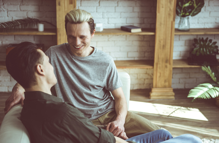 Toned portrait of handsome young man sitting on couch with dark-haired friend and holding his hand. Wall unit with shelves on backgroundの写真素材