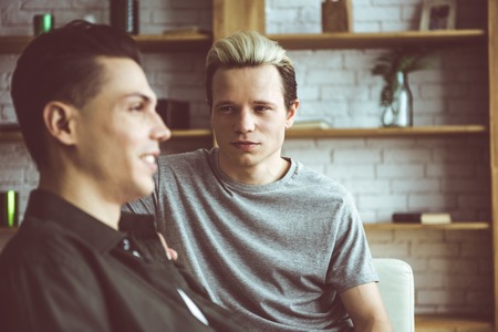 Toned portrait of handsome guy sitting on couch and gazing at dark-haired smiling friend. Focus on blond gentlemanの写真素材