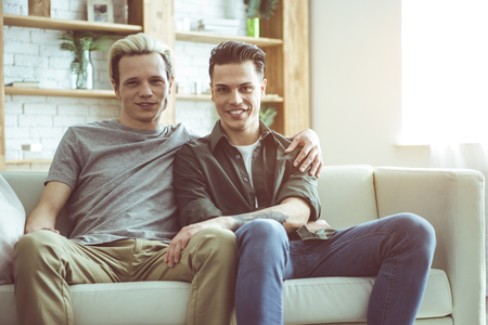 We are happy. Toned portrait of gay couple sitting on white couch and cuddling. Guys looking at camera with happy smilesの写真素材