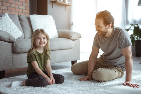 I like playing with my daddy. Full length portrait of cute blonde girl sitting on soft carpet near her father in apartment. She is looking at camera and laughingの写真素材