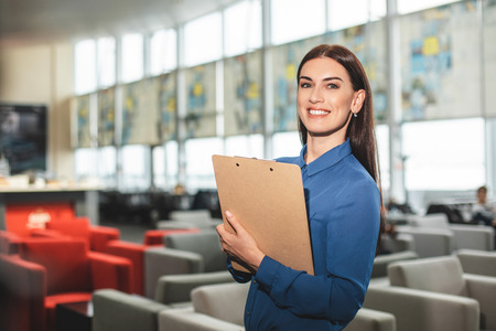 Waist up portrait of cheerful businesswoman using folder and looking at camera with bright smile. Copy space on left sideの写真素材