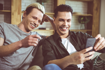 This is hilarious. Toned portrait of gay couple sitting on couch at home and watching funny videos on cellphoneの写真素材