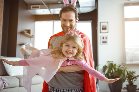Dreams come true. Portrait of happy little kid is flying on dad hands. She is wearing robe and smiling. Daughter is looking at camera with excitementの写真素材