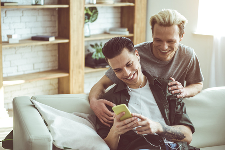 Toned portrait of dark-haired guy in headphones sitting on couch and holding cellphone while his boyfriend hugging him from behind. They looking at phone screen and smilingの写真素材