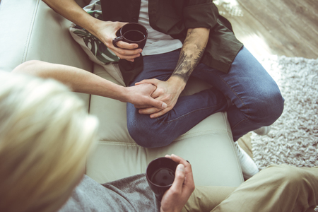 Romantic morning. Top view portrait of gay couple holding hands while sitting on couch with cups of coffeeの写真素材