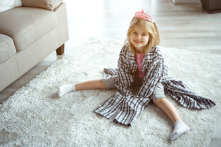 Playful kid is having fun at home. Little princess is wearing large clothing of her daddy while sitting on floor. She is looking at camera and laughingの写真素材