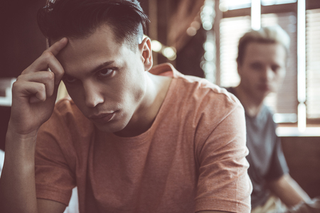Close up portrait of dark-haired handsome guy looking away with unhappy expression and touching forehead. His boyfriend sitting behind on blurred backgroundの写真素材