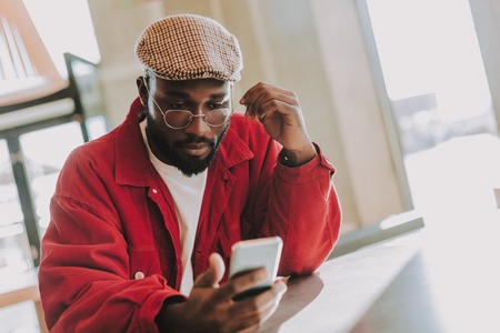 Serious glance. Young bearded man sitting in the cafe alone and looking attentively at the screen of...