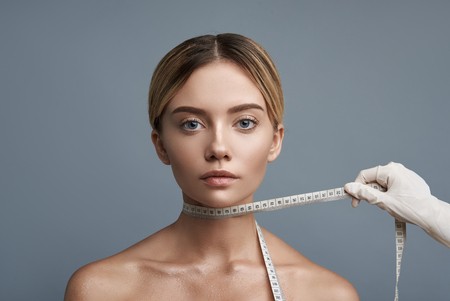 Measuring tape. Calm young woman sitting against the grey background and looking at you while having measuring tape on her neckの写真素材