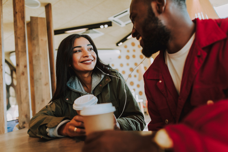Sharing earphones. Cheerful lovely young couple sharing earphones and listening to pleasant music while sitting together and drinking coffeeの写真素材