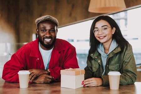 Positive young couple sitting at the table and smiling happily while having cups of coffee and a gift box in front of themの写真素材