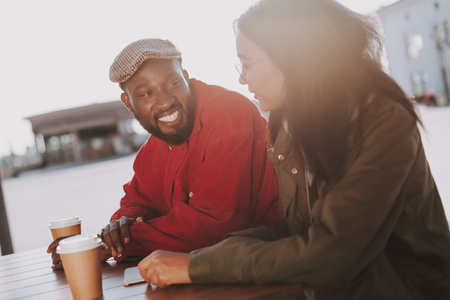 Positive young man kindly smiling to his girlfriend while sitting at the table outdoors with herの写真素材