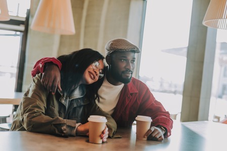 Hugging couple. Peaceful young lady feeling relaxed while putting her head on the shoulder of her handsome cheerful boyfriend and sitting in the cafe with himの写真素材