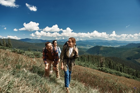 Cheerful man and two women hiking in mountains. They are travelling with rucksacks together along path up to topの写真素材