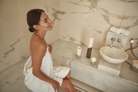 Happy young woman smiling and looking happy while sitting in hammam and touching the towel on her bodyの写真素材