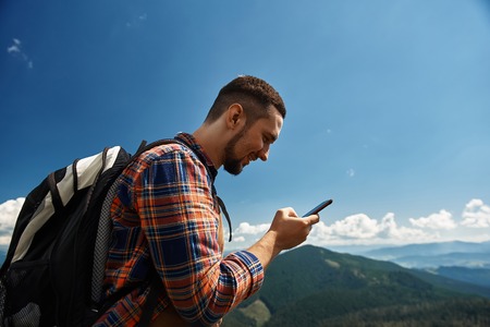 Merry man standing on top among high mountains. He carrying rucksack and holding mobile phoneの写真素材