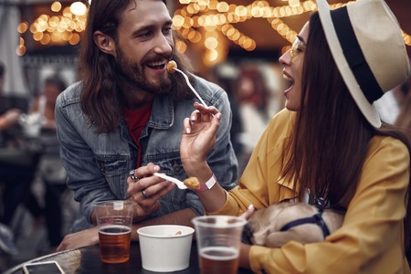 Portrait of hipster young lady in hat letting man taste food in outdoor cafe.