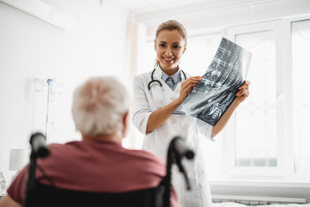 Portrait of young lady in white lab coat holding x-ray image and pointing at it while patient sitting in wheelchair. Focus on womanの写真素材