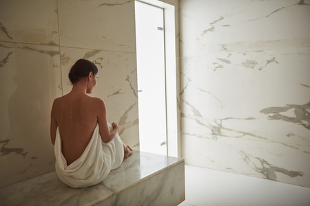 Peaceful dark haired lady covered in white towel and showing her back after pleasant hammam...