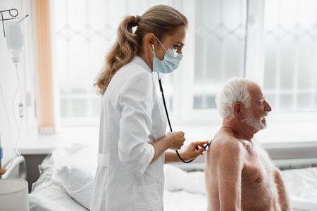 Side view portrait of shirtless bearded gentleman sitting on hospital bed and taking long breath during medical examinationの写真素材