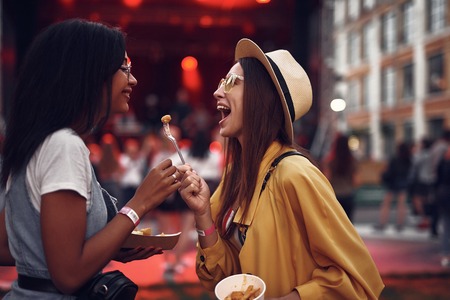 Taste this. Side view portrait of lovely girl in hat holding for with french fry while her friend looking at it and smilingの写真素材