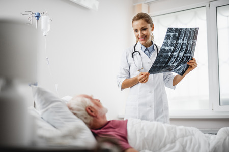 Good news. Portrait of beautiful young lady in white lab coat sharing radiography results with old man. Gentleman lying in hospital bedの写真素材