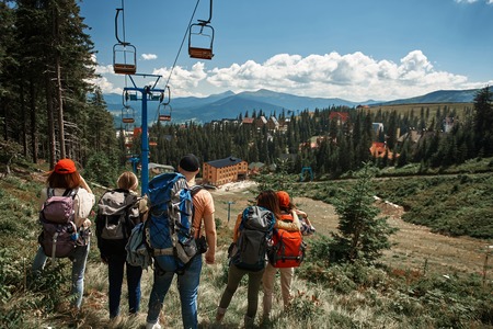 Four women and one man travelling through mountains with rucksacks. They standing with focus on back near chair lift and staring forward at village downhillの写真素材