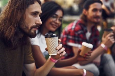 Calm bearded young man sitting next to his friends and thoughtfully looking into the distance while drinking coffeeの写真素材
