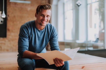 Waist up portrait of young happy designer is siting with new interesting project in his hands and smilingの写真素材