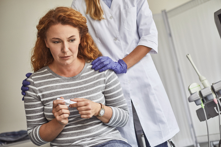 Waist up portrait of unhappy red-haired woman holding pregnancy test with negative result. Supportive doctor comforting lady and touching her shouldersの写真素材