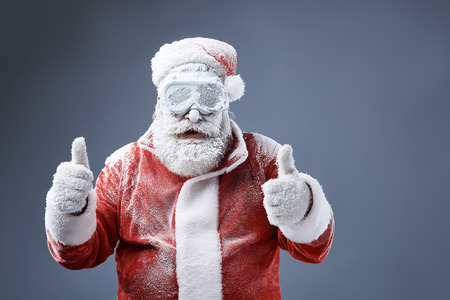 Portrait of bearded old man in Santa costume covered with snow demonstrating approval gesture. Isolated on gray-blue backgroundの写真素材
