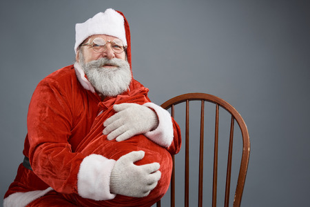 Waist up portrait of bearded old man in Santa costume hugging bag with christmas presents. Isolated on gray backgroundの写真素材