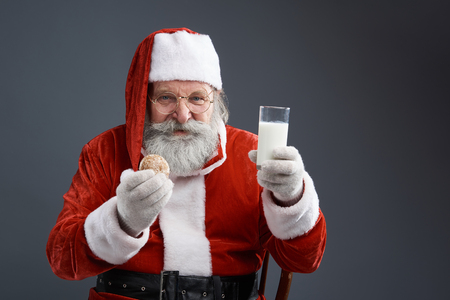 Waist up portrait of bearded old man in Santa costume sitting on chair and demonstrating cookie with drink. Isolated on gray backgroundの写真素材