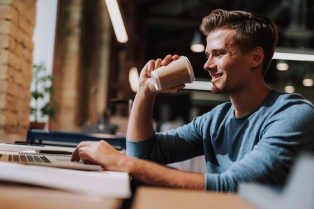 Waist up portrait of smiling man sitting in coworking space and working with laptop while holding hot drinkの写真素材