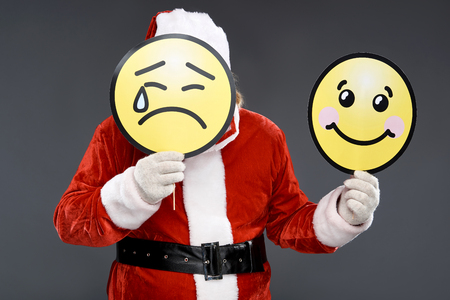 Waist up portrait of old man in Santa costume hiding under emoticon icon with sad face while demonstrating carton sign of happiness. Isolated on gray backgroundの写真素材